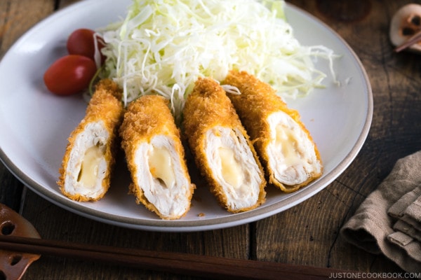 A white ceramic plate containing chicken cheese katsu served with shredded cabbage and cherry tomato.