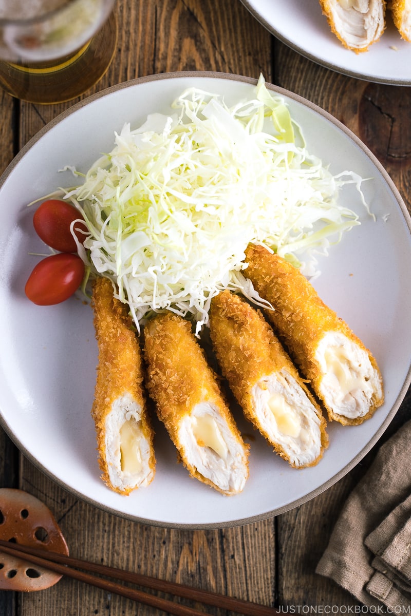 A white ceramic plate containing chicken cheese katsu served with shredded cabbage and cherry tomato.