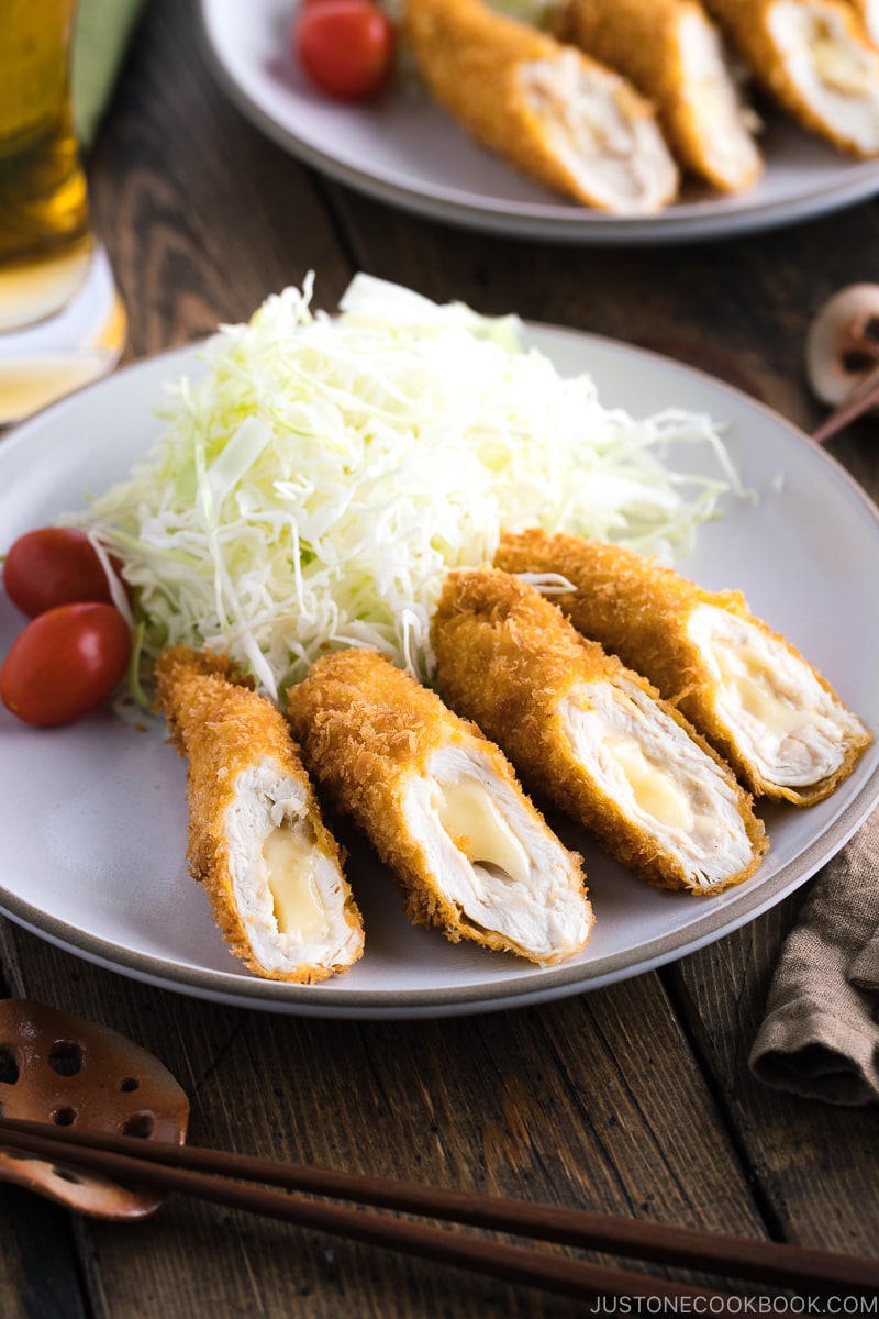 A white ceramic plate containing chicken cheese katsu served with shredded cabbage and cherry tomato.