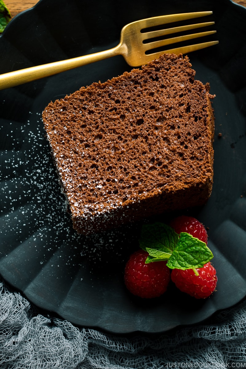 A slice of chocolate chiffon cake and raspberries are served on a black matte plate.