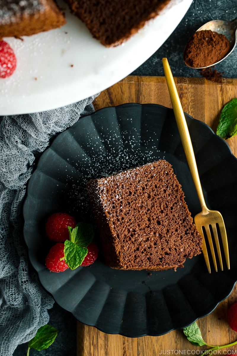 A slice of chocolate chiffon cake and raspberries are served on a black matte plate.