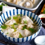 a large bowl containing shrimp and wonton soup along with the condiments.