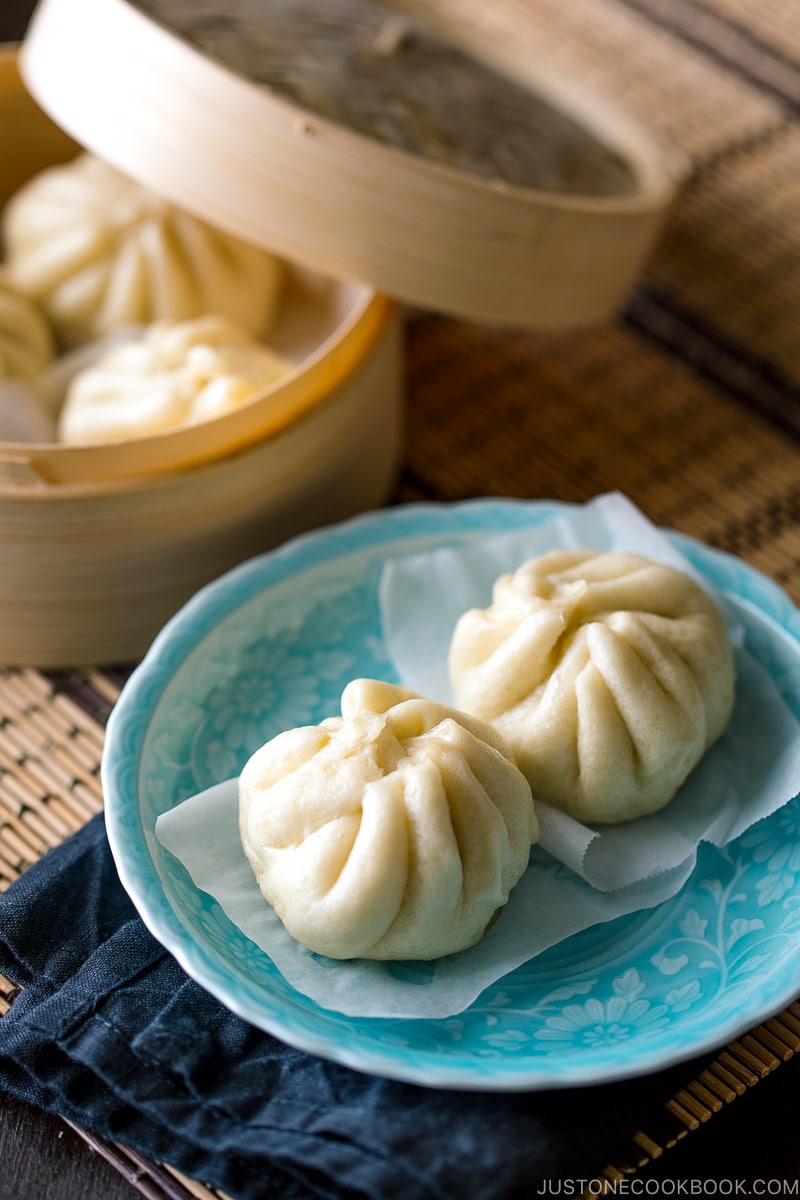 A mini bamboo steamer and a blue dish containing Steamed Pork Buns (Nikuman).