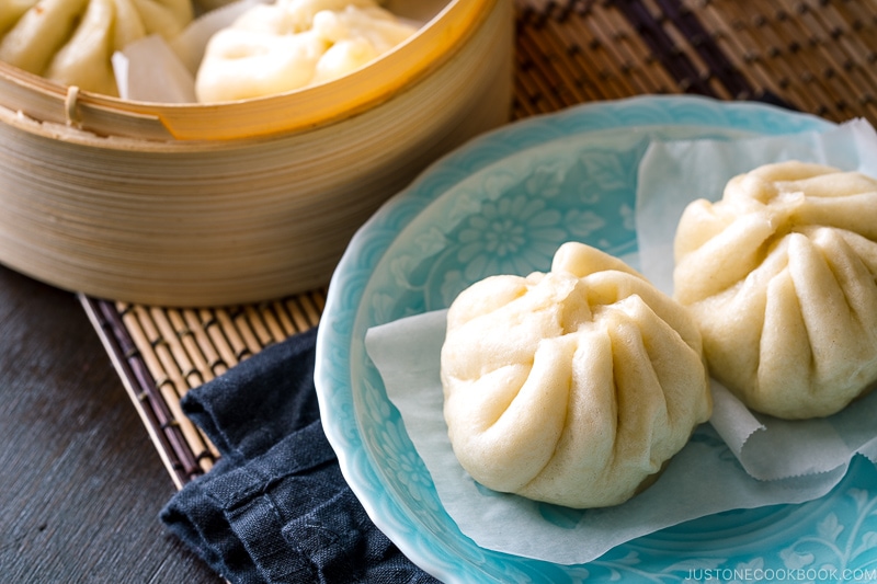 A mini bamboo steamer and a blue dish containing Steamed Pork Buns (Nikuman).