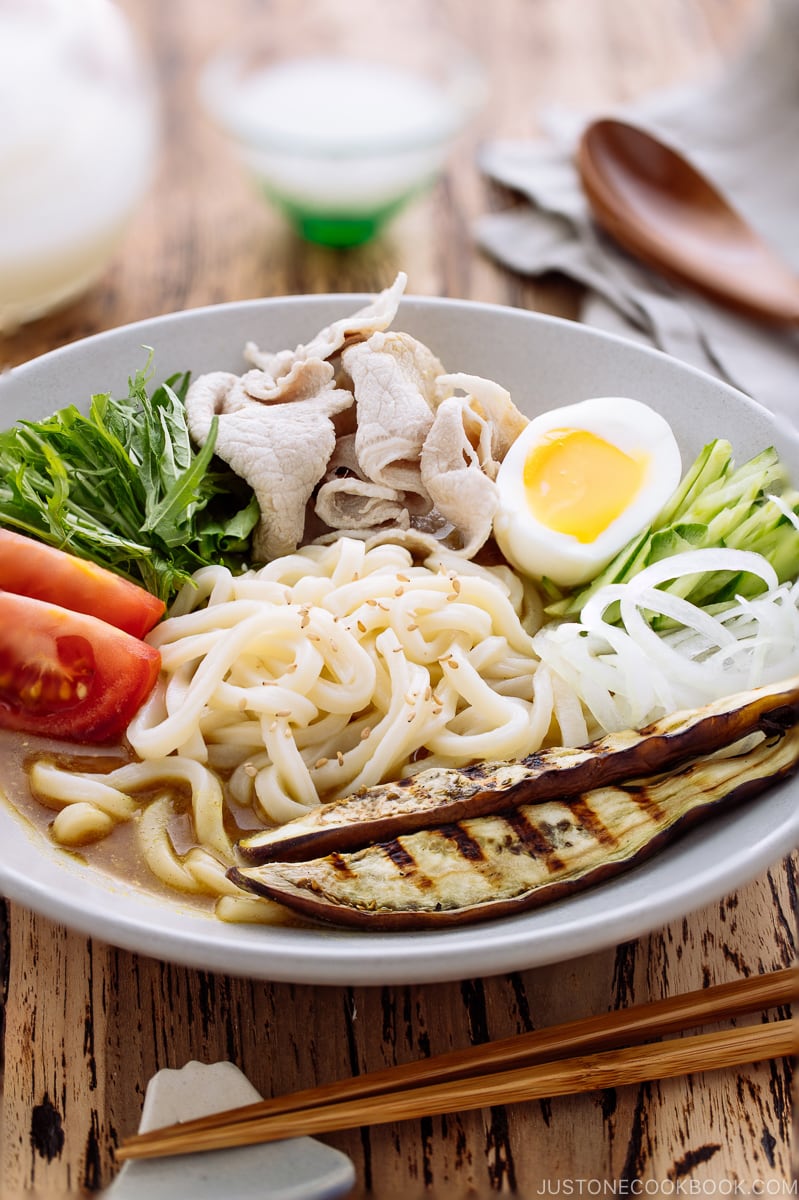 A white bowl containing cold curry udon with sliced pork, boiled egg, and fresh vegetables.