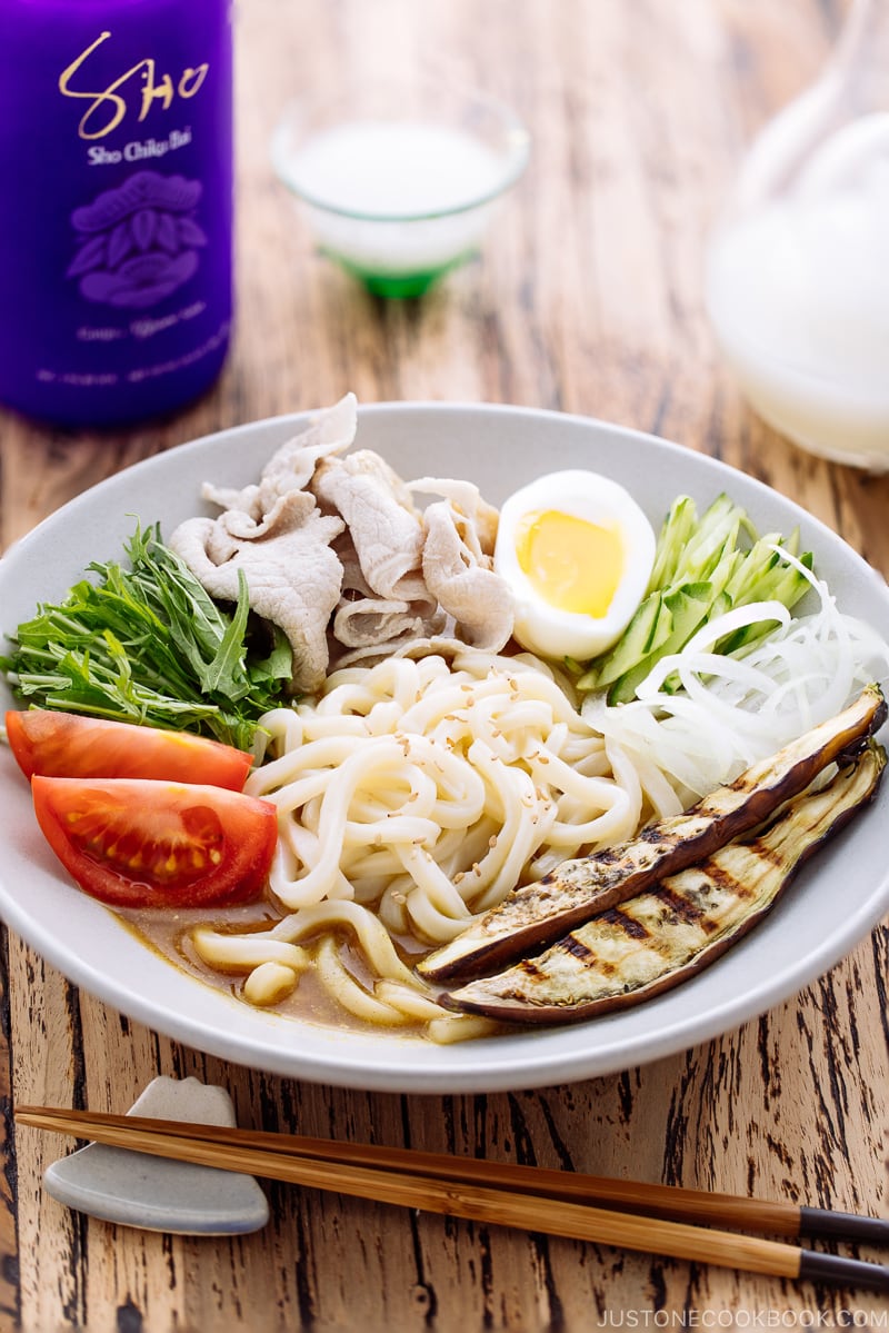 A white bowl containing cold curry udon with sliced pork, boiled egg, and fresh vegetables.