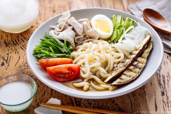 A white bowl containing cold curry udon with sliced pork, boiled egg, and fresh vegetables.