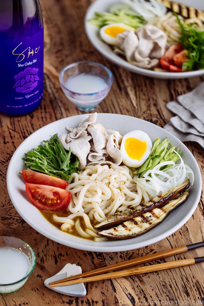 A white bowl containing cold curry udon with sliced pork, boiled egg, and fresh vegetables.