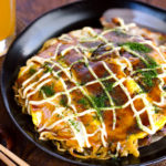 A black plate containing Hiroshima-style OKonomiyaki topped with the savory sauce and Japanese mayo.