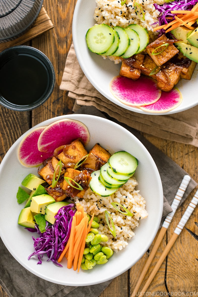 A white bowls containing savory pan-fried tofu, cucumber, avocado, edamame, carrot, red cabbage, and watermelon radish over brown rice.