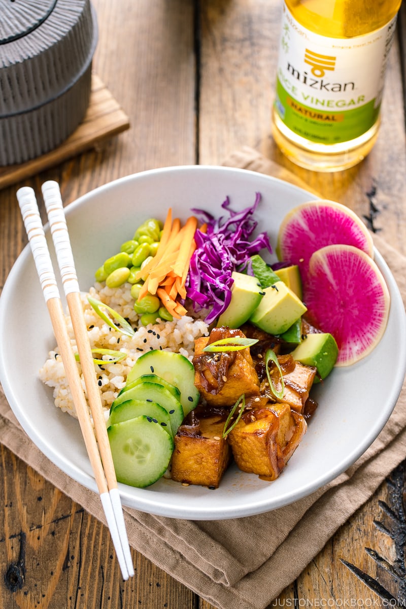 A white bowls containing savory pan-fried tofu, cucumber, avocado, edamame, carrot, red cabbage, and watermelon radish over brown rice.