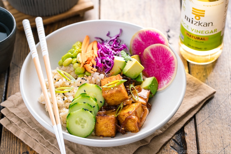 A white bowls containing savory pan-fried tofu, cucumber, avocado, edamame, carrot, red cabbage, and watermelon radish over brown rice.