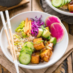 A white bowls containing savory pan-fried tofu, cucumber, avocado, edamame, carrot, red cabbage, and watermelon radish over brown rice.