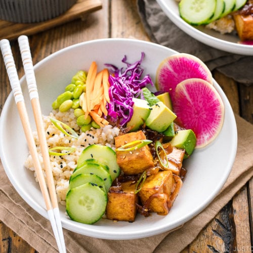 A white bowls containing savory pan-fried tofu, cucumber, avocado, edamame, carrot, red cabbage, and watermelon radish over brown rice.