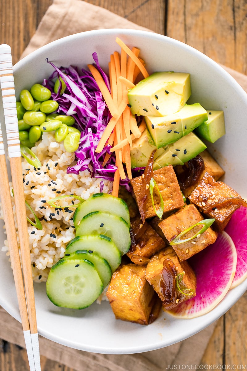 A white bowls containing savory pan-fried tofu, cucumber, avocado, edamame, carrot, red cabbage, and watermelon radish over brown rice.