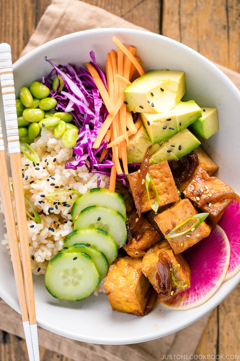 Un bol blanc contenant du tofu poêlé salé, du concombre, de l'avocat, de l'edamame, de la carotte, du chou rouge et du radis pastèque sur du riz brun.