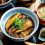 A Japanese rice bowl containing Soy-Glazed Eggplant Donburi topped with julienned shiso and toasted sesame seeds.