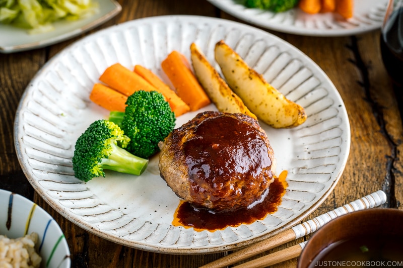 A white plate containing Japanese Hamburger Steak (Hambagu), sautéed carrot, broccoli, and baked potato wedges.