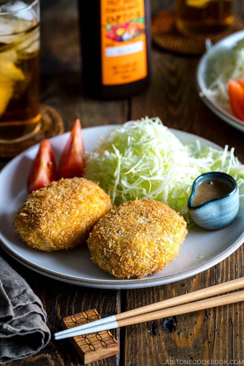 A white plate containing Korokke served with Tonkatsu sauce and shredded cabbage on the side.