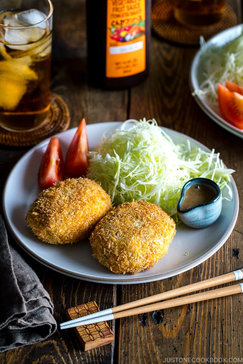 A white plate containing Korokke served with Tonkatsu sauce and shredded cabbage on the side.