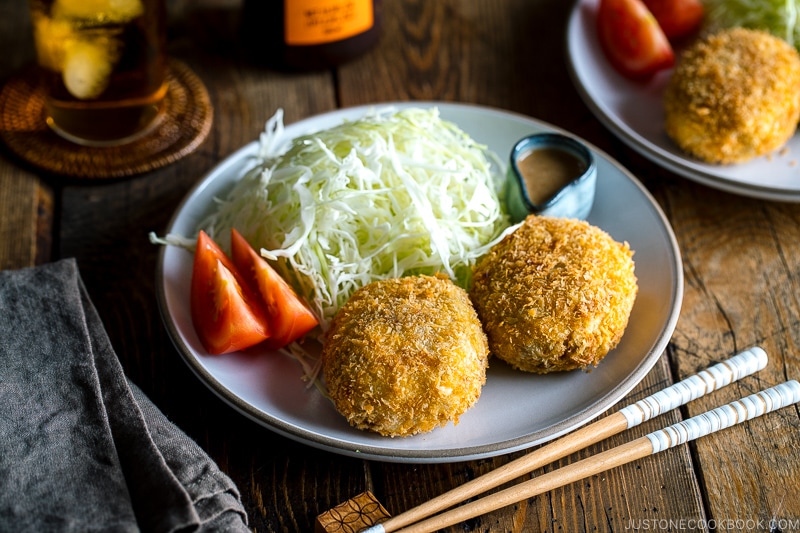 A white plate containing Korokke served with Tonkatsu sauce and shredded cabbage on the side.