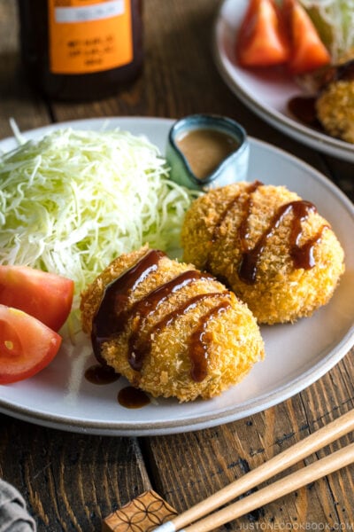 A white plate containing Japanese Croquette (Korokke) served with Tonkatsu sauce and shredded cabbage on the side.
