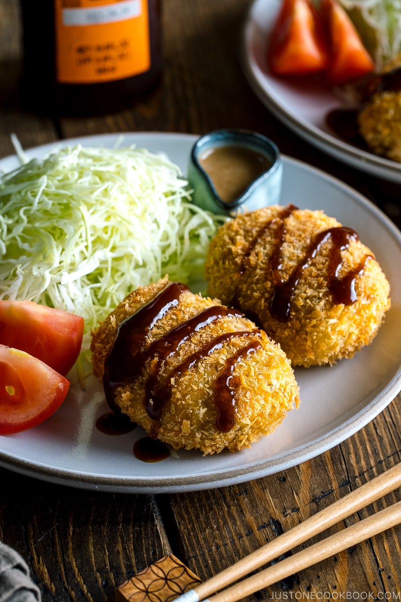 A white plate containing Japanese Croquette (Korokke) served with Tonkatsu sauce and shredded cabbage on the side.