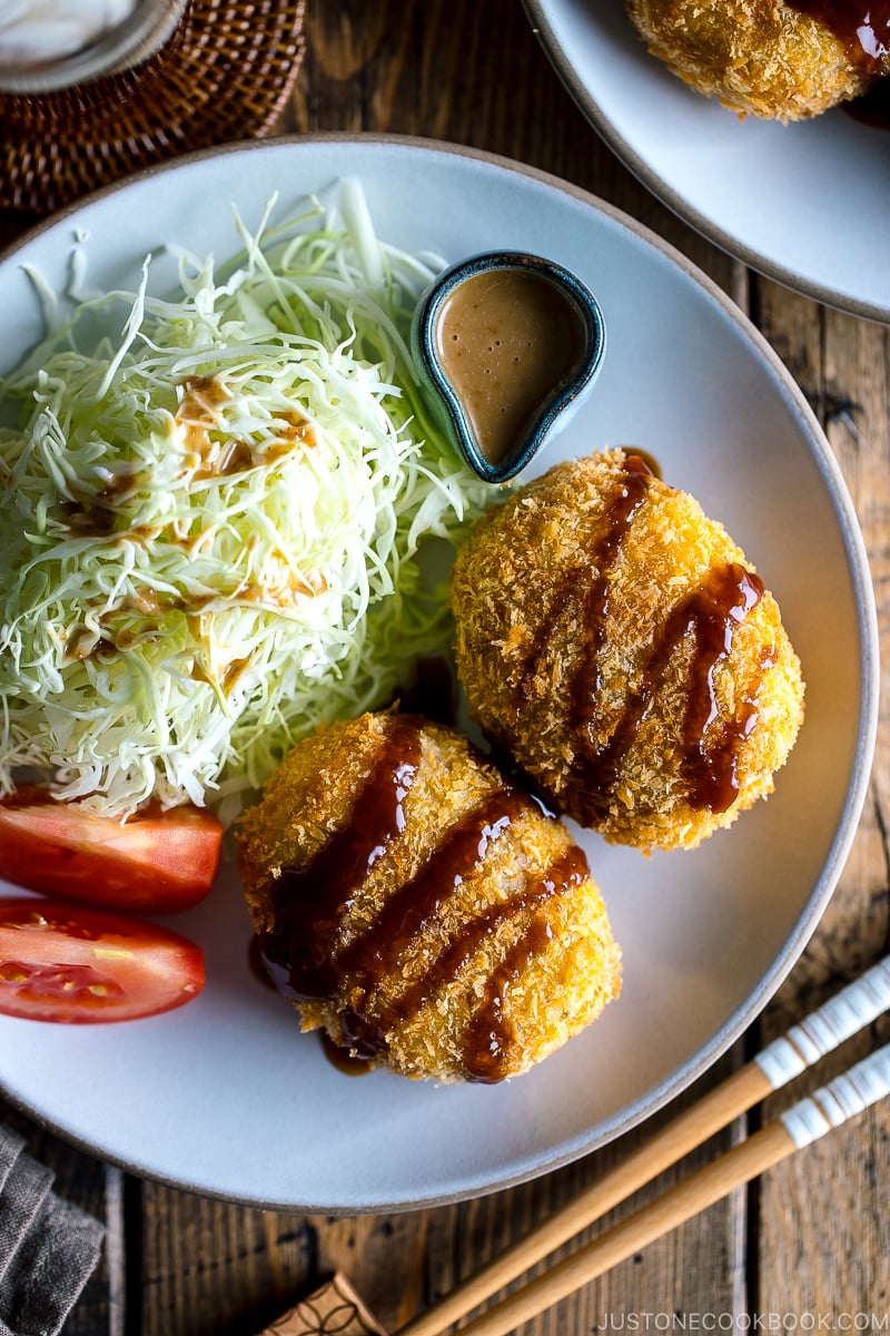 A white plate containing Korokke served with Tonkatsu sauce and shredded cabbage on the side.