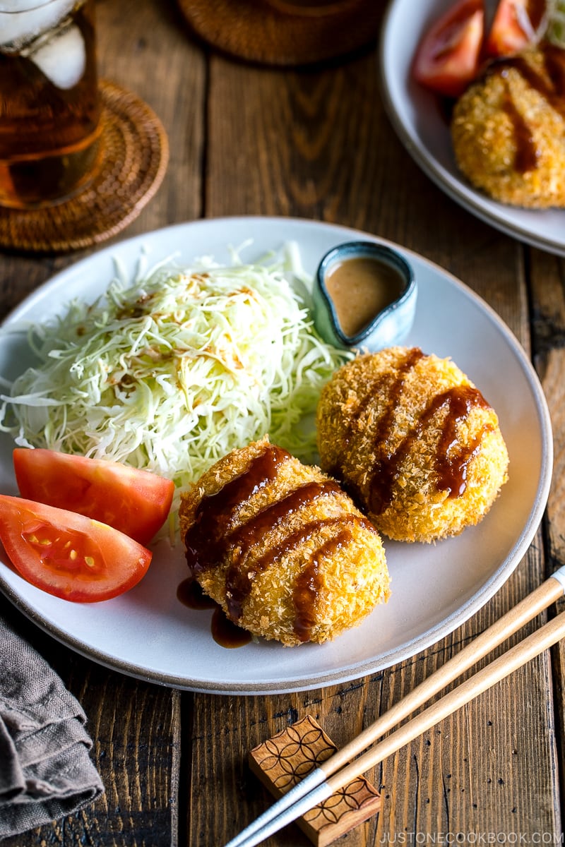 A white plate containing Japanese Croquette (Korokke) served with Tonkatsu sauce and shredded cabbage on the side.