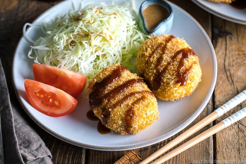 A white plate containing Japanese Croquette (Korokke) served with Tonkatsu sauce and shredded cabbage on the side.