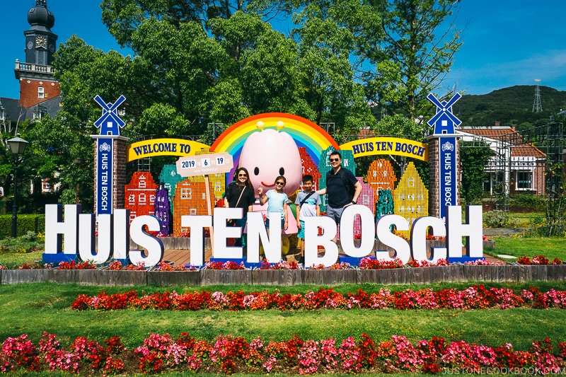 a family standing behind Huis Ten Bosch sign in a flowerbed