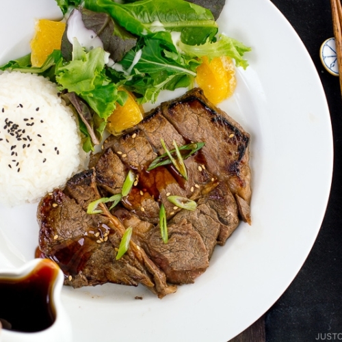 A white plate containing beef teriyaki served with steamed rice and salad.