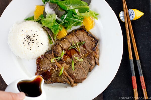 A white plate containing beef teriyaki served with steamed rice and salad.