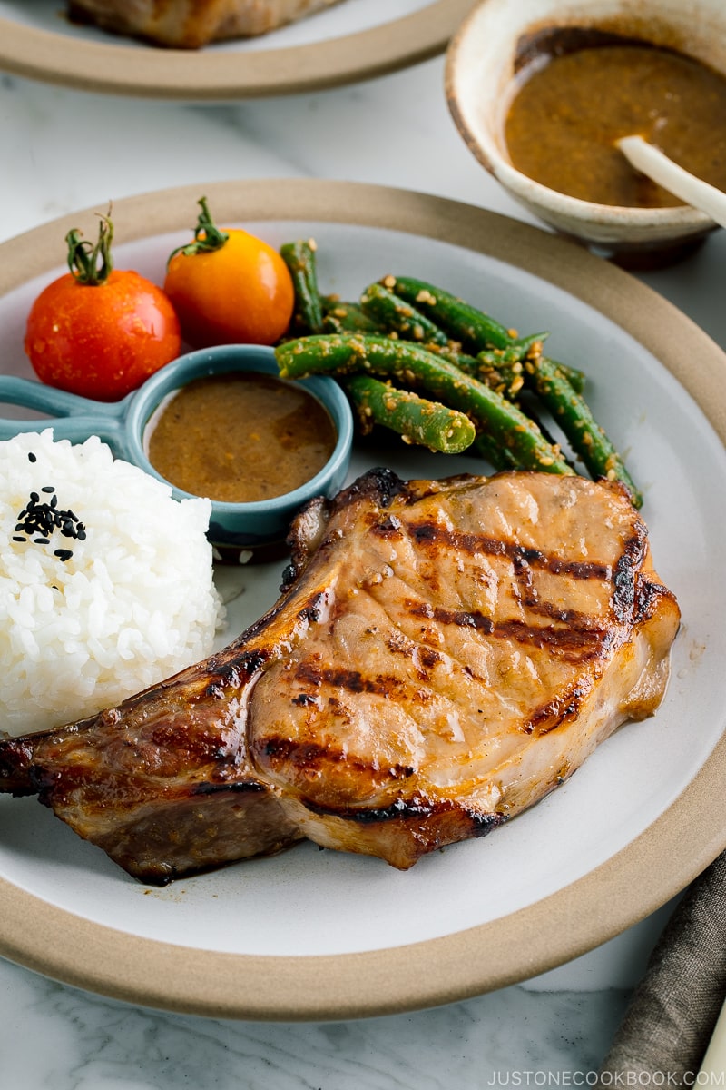 A white plate containing the kurobuta pork chop with yuzu kosho flavored miso sauce, along with rice, green bean gomaae, and tomatoes..
