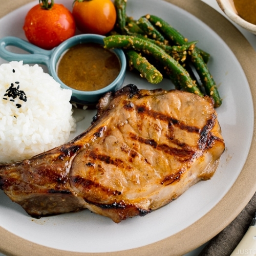 A white plate containing the kurobuta pork chop with yuzu kosho flavored miso sauce, along with rice, green bean gomaae, and tomatoes..