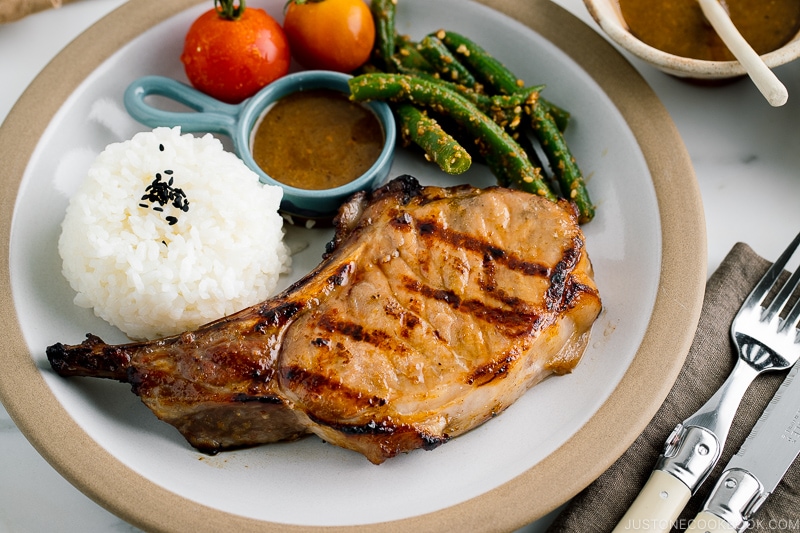 A white plate containing the kurobuta pork chop with yuzu kosho flavored miso sauce, along with rice, green bean gomaae, and tomatoes..