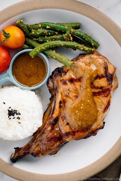 A white plate containing the kurobuta pork chop with yuzu kosho flavored miso sauce, along with rice, green bean gomaae, and tomatoes..