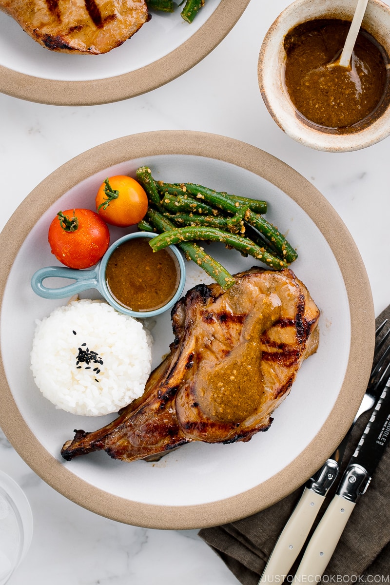 A white plate containing the kurobuta pork chop with yuzu kosho flavored miso sauce, along with rice, green bean gomaae, and tomatoes..