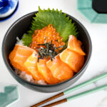 A black bowl containing a bed of rice topped with sashimi grade salmon, ikura, and shredded nori.