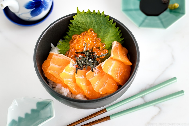 A black bowl containing a bed of rice topped with sashimi grade salmon, ikura, and shredded nori.