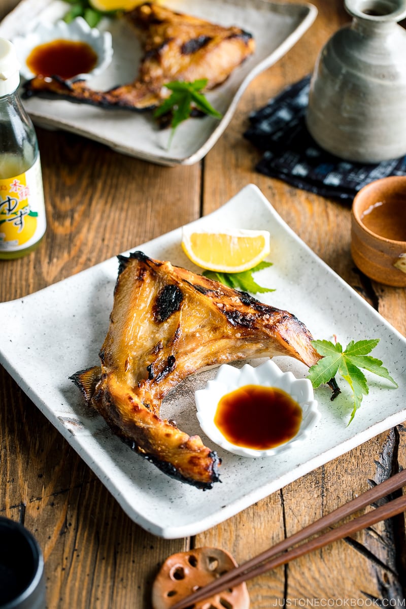 A white Japanese ceramic plate containing Hamachi Kama (Grilled Yellowtail Collar) along with a wedge of lemon and yuzu-soy dipping sauce.