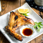 A white Japanese ceramic plate containing Hamachi Kama (Grilled Yellowtail Collar) along with a wedge of lemon and yuzu-soy dipping sauce.