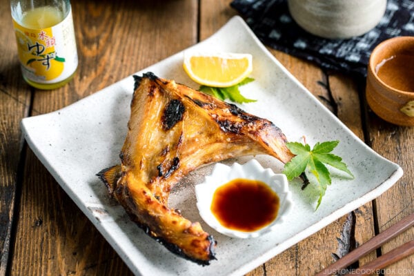 A white Japanese ceramic plate containing Hamachi Kama (Grilled Yellowtail Collar) along with a wedge of lemon and yuzu-soy dipping sauce.