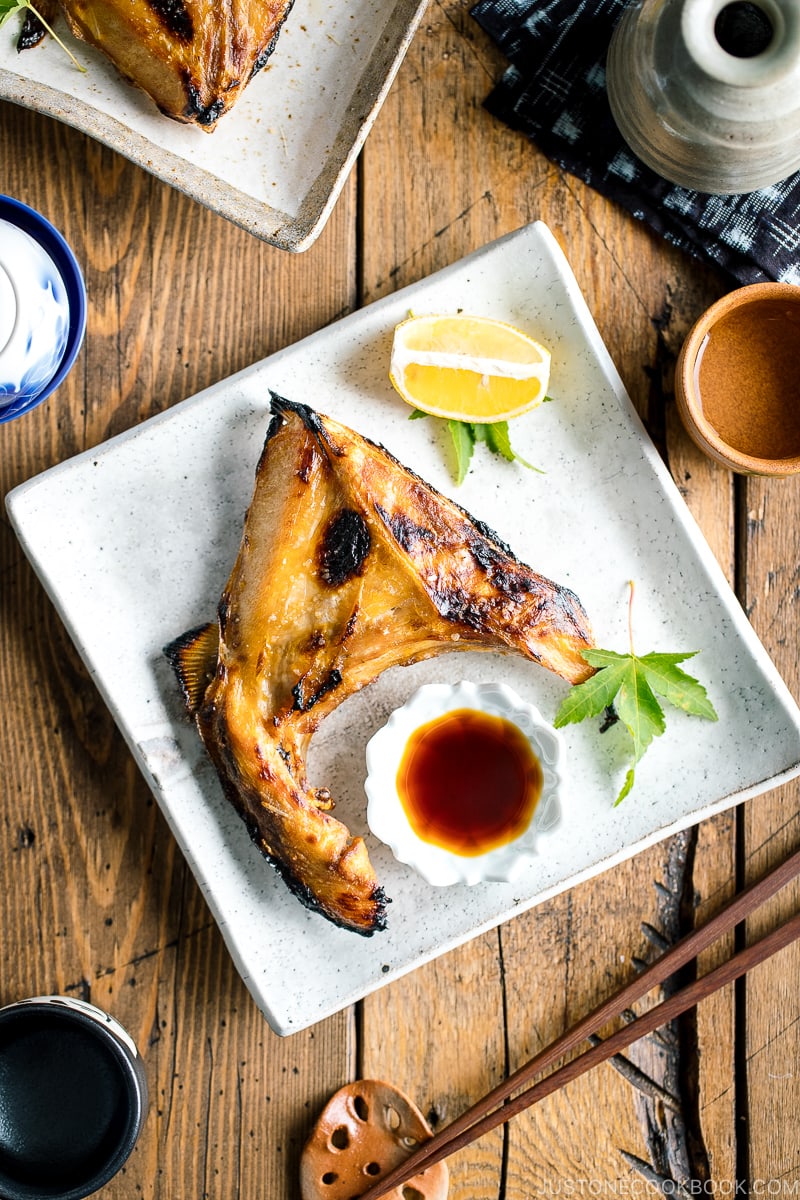 A white Japanese ceramic plate containing Hamachi Kama (Grilled Yellowtail Collar) along with a wedge of lemon and yuzu-soy dipping sauce.