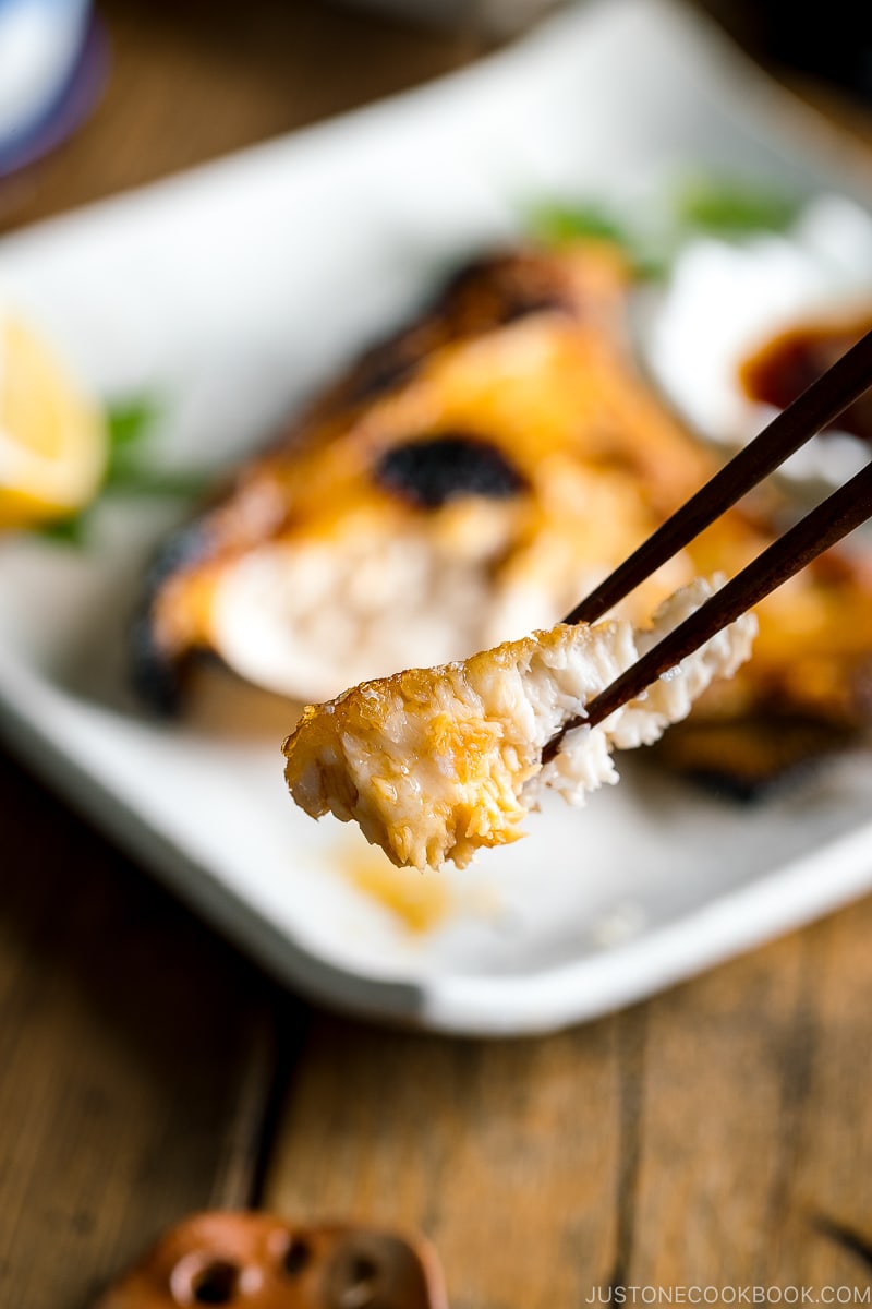 A white Japanese ceramic plate containing Hamachi Kama (Grilled Yellowtail Collar) along with a wedge of lemon and yuzu-soy dipping sauce.