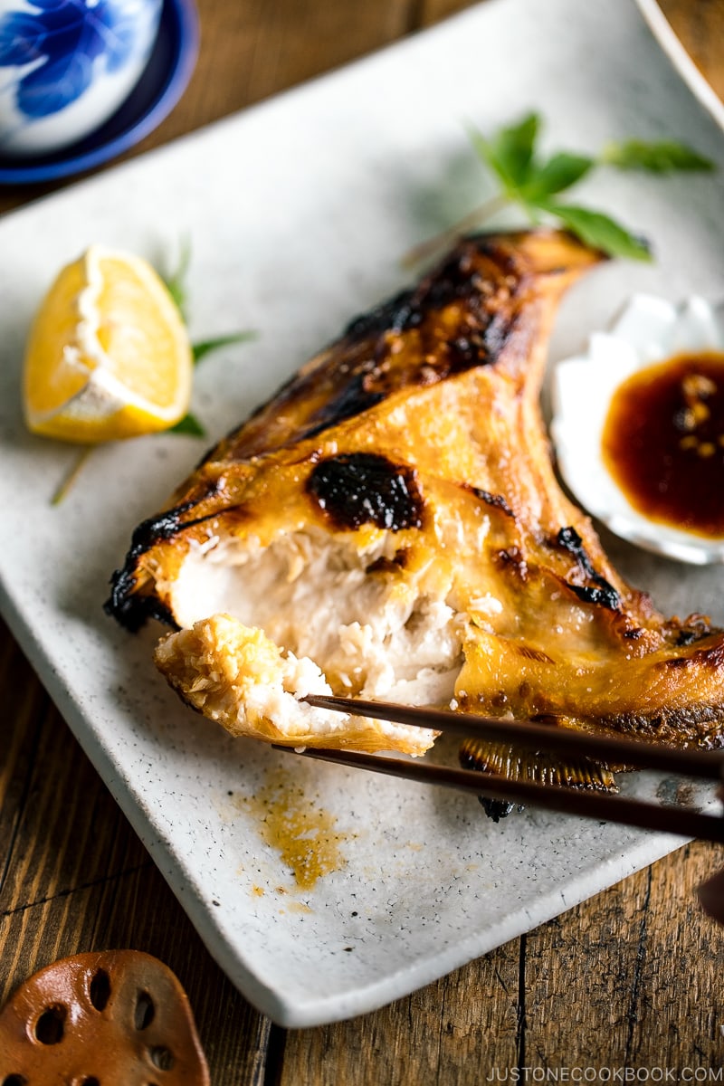 A white Japanese ceramic plate containing Hamachi Kama (Grilled Yellowtail Collar) along with a wedge of lemon and yuzu-soy dipping sauce.