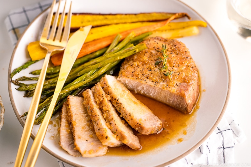 A white plate containing honey garlic pork chops, roasted carrot, and asparagus.