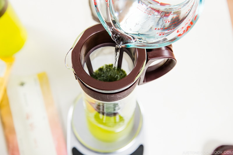 water being poured into a cold brew pitcher with green tea leaves