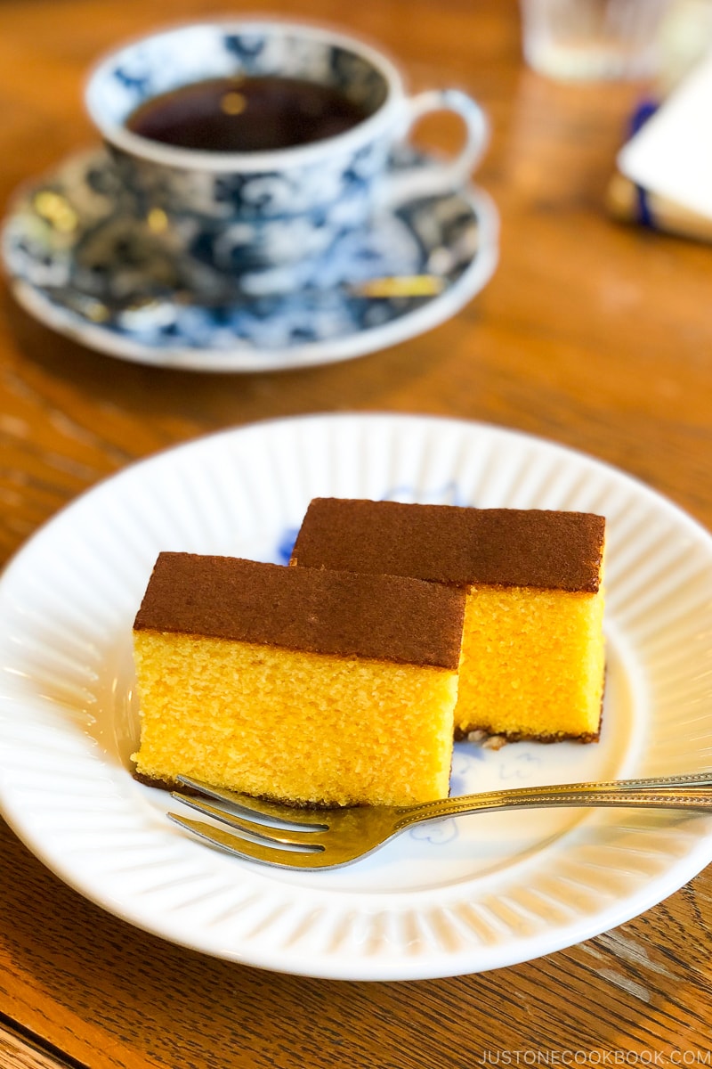 castella cake on a white plate next to a cup of coffee