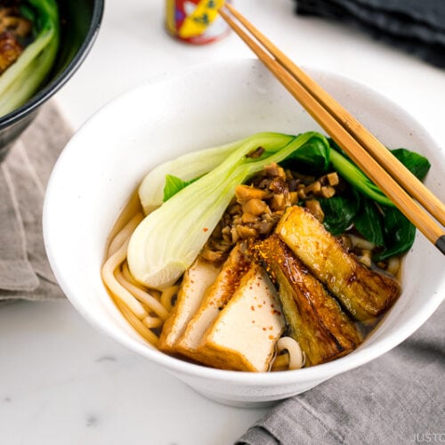A white bowl containing Vegetarian Udon Noodle Soup topped with bok choy, fried tofu, minced mushroom, and crispy eggplant.